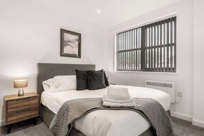 A bedroom with a bed, a nightstand, a framed picture, a window with blinds, and a radiator. - Modern 1 Bedroom Apartment in Leeds - Sleeps 3 (Leeds)