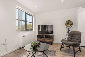 A modern living room with a large TV, a comfortable chair, a small table with a plant, and a radiator. - Modern 1 Bedroom Apartment in Leeds - Sleeps 3 (Leeds)