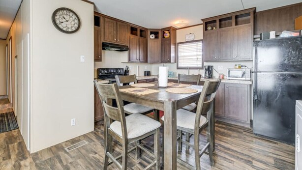 Une cuisine de taille réduite avec des meubles de rangement en bois, une table à manger avec des chaises, une horloge au mur et un réfrigérateur.