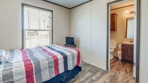 Une chambre à coucher avec un lit, une fenêtre avec des stores, un téléviseur et une salle de bain visible à travers une porte ouverte.