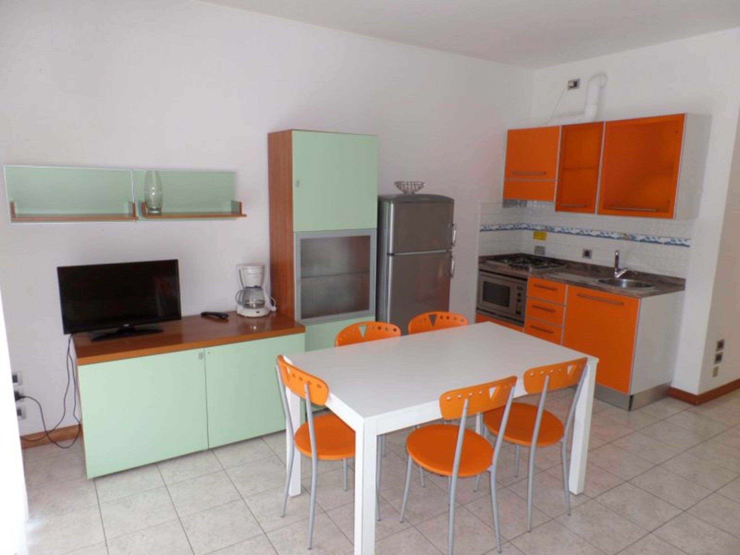 Een kleine keuken met een eettafel, stoelen, een koelkast, een fornuis en een televisie.
