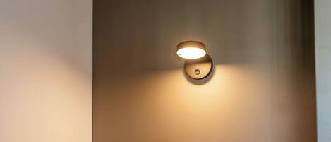 A neatly made bed with a headboard against a plain wall, a wall-mounted light fixture, and a small circular object on the wall.
