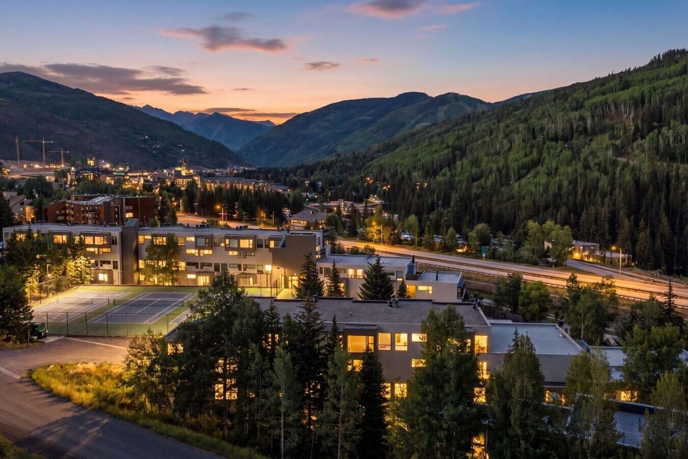 A modern building complex with multiple floors, surrounded by a forested area and mountains in the background.
