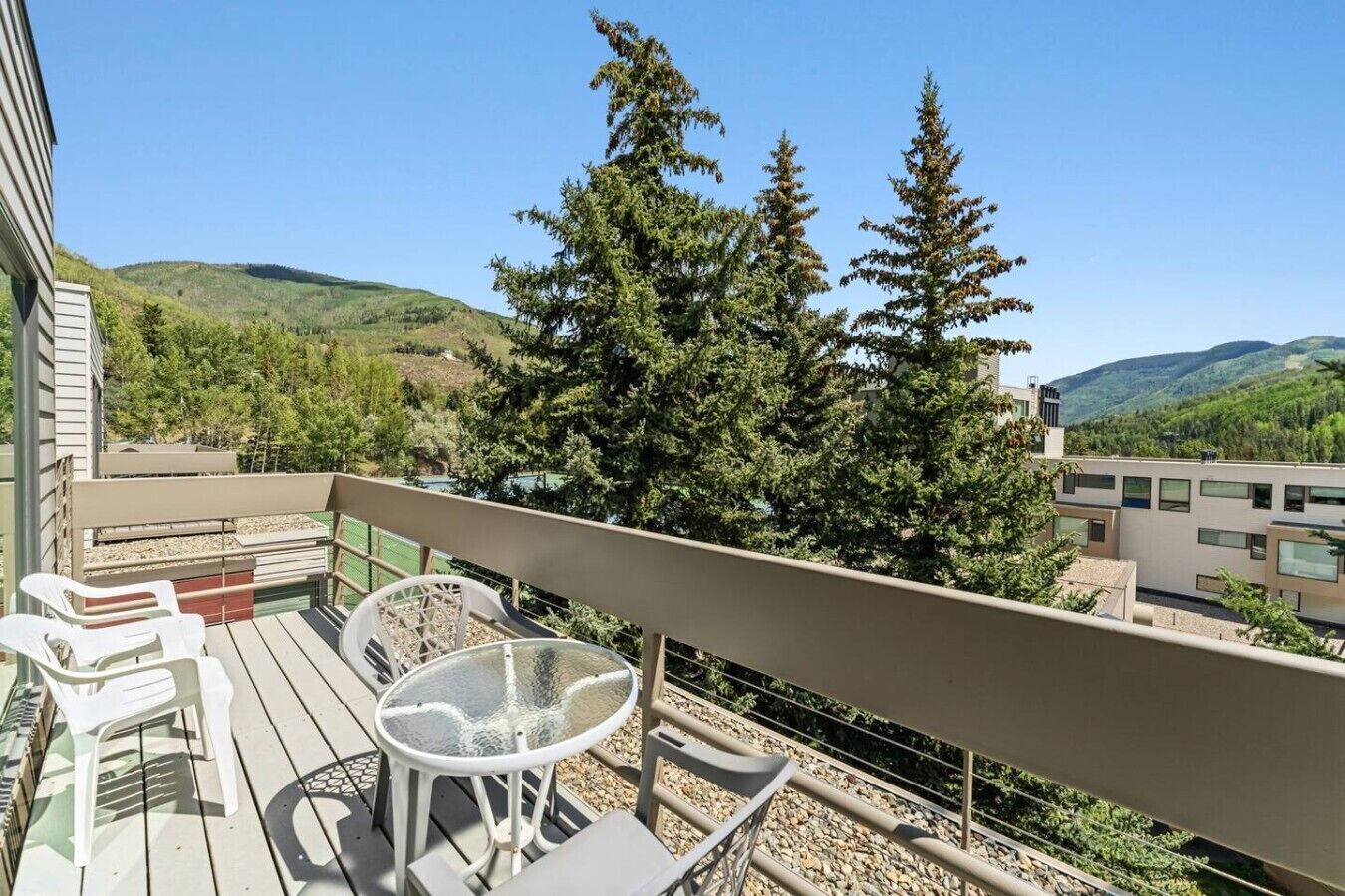 A balcony with outdoor furniture, a glass table, and a view of trees and hills.