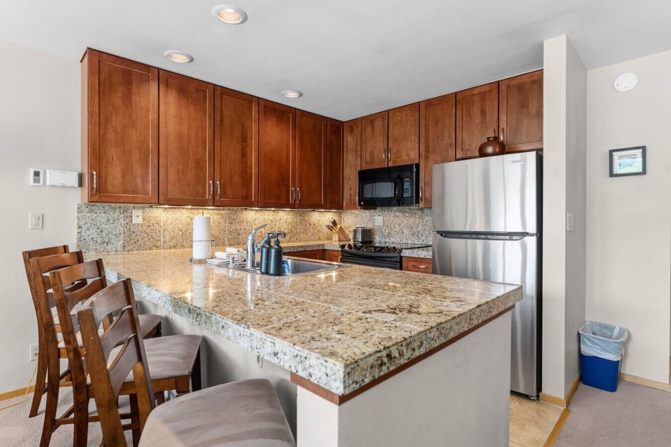 A kitchen with wooden cabinets, a stainless steel refrigerator, a microwave, granite countertops, and bar stools.