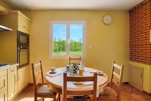 Un coin repas comprenant une table ronde, des chaises, une horloge et une fenêtre donnant sur des arbres.