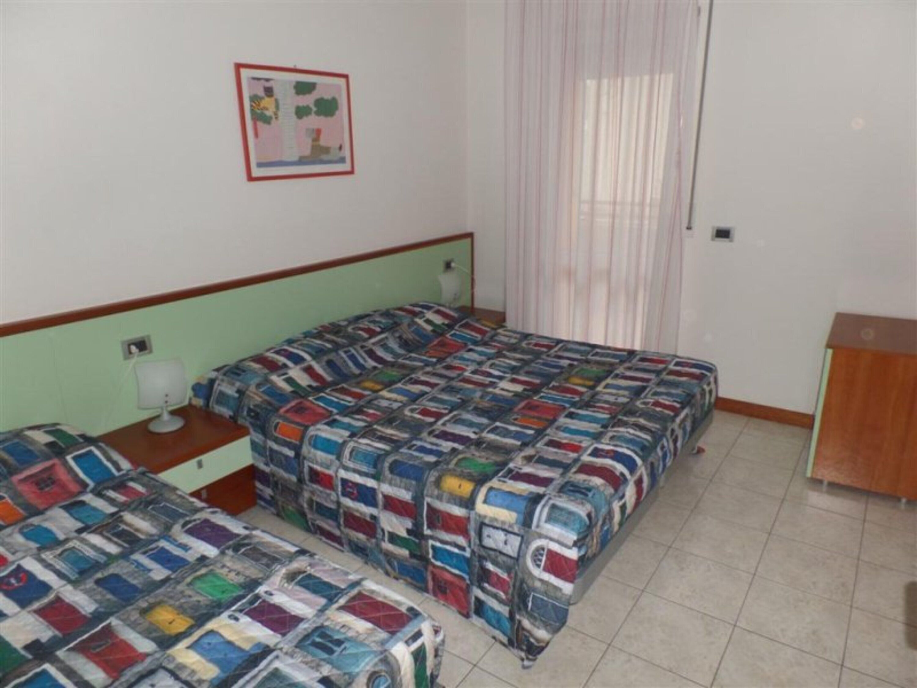 A bedroom with two beds, a bedside table, a lamp, a picture on the wall, and a window with curtains.