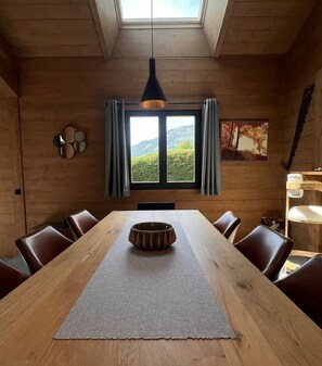 A dining area with a long wooden table, leather chairs, a central decorative bowl, a skylight, and a window with curtains. - PRAZ SUR ARLY Chalet, 5 minutes from MEGEVE—Come experience it for yourself (PRAZ SUR ARLY,)