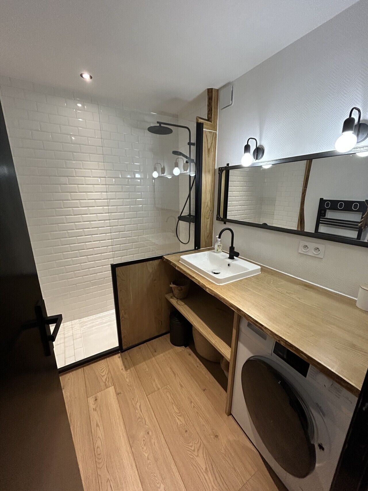 Een moderne badkamer met een witte betegelde douche, een houten wastafelmeubel met een wastafel, en een ingebouwde wasmachine.