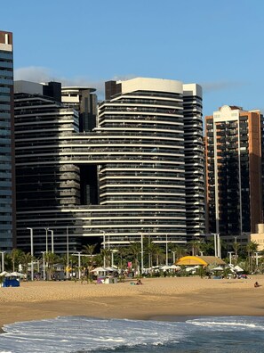 Ein modernes Hotel mit mehreren Etagen und Balkonen, nahe einem Strand mit Palmen und Sonnenschirmen.
