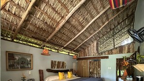 Een kamer met rieten dak, een bed met een blauwe deken, een houten stoel, een klein tafeltje met flessen en een lijst met een foto aan de muur.