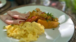 Een bord eten met roerei, ham en een groenterecht, mogelijk curry, op een tafel met een geweven placemat.