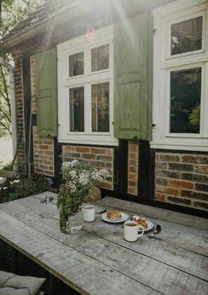 Ein rustikaler Holztisch mit Blumen in einer Vase, zwei Teller mit Speisen und einer Tasse, vor einer Backsteinwand mit grünen Fensterläden.