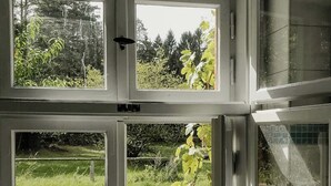 Huoneessa on suuri ikkuna, joka on jaettu neljään osaan, ja siitä avautuu näkymä puutarhaan, jossa on puinen aita ja runsaasti vihreää kasvillisuutta.