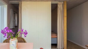 Una habitación con piso de madera, una mesa con un florero, una silla, una cama con cortina a rayas y una puerta que da a otra habitación.
