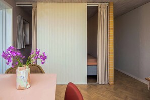 Ein Zimmer mit Holzboden, einem Tisch mit Blumen, einem Stuhl, einem Bett mit einem Streifenrollo und einer Tür zu einem weiteren Raum.