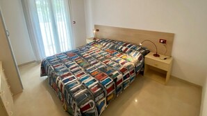 A bedroom with a bed, a bedside table, a lamp, a nightstand, and a window with curtains. - Modern Stay in a Central Area in Bibione (San Michele al Tagliamento)