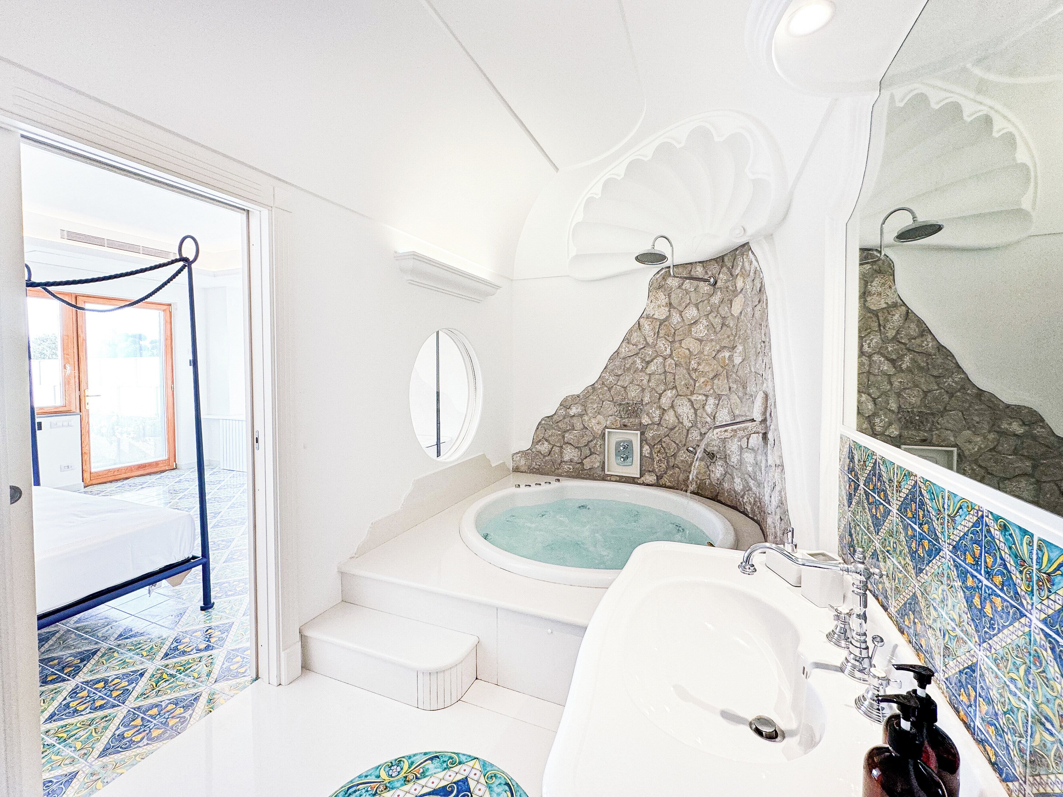 Un baño con bañera, lavabo y espejo. Se observa una pared de piedra y una puerta que da a otra habitación.