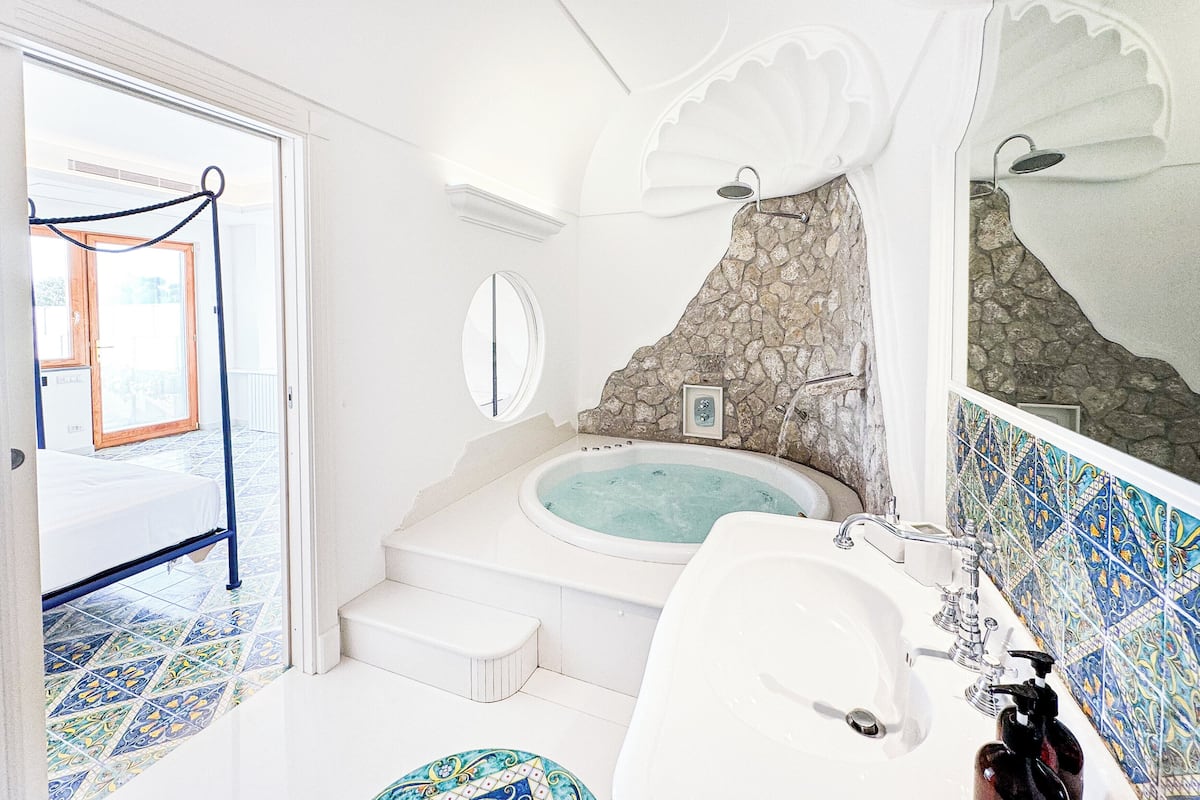 Un baño con bañera, lavabo y espejo. Se observa una pared de piedra y una puerta que da a otra habitación.