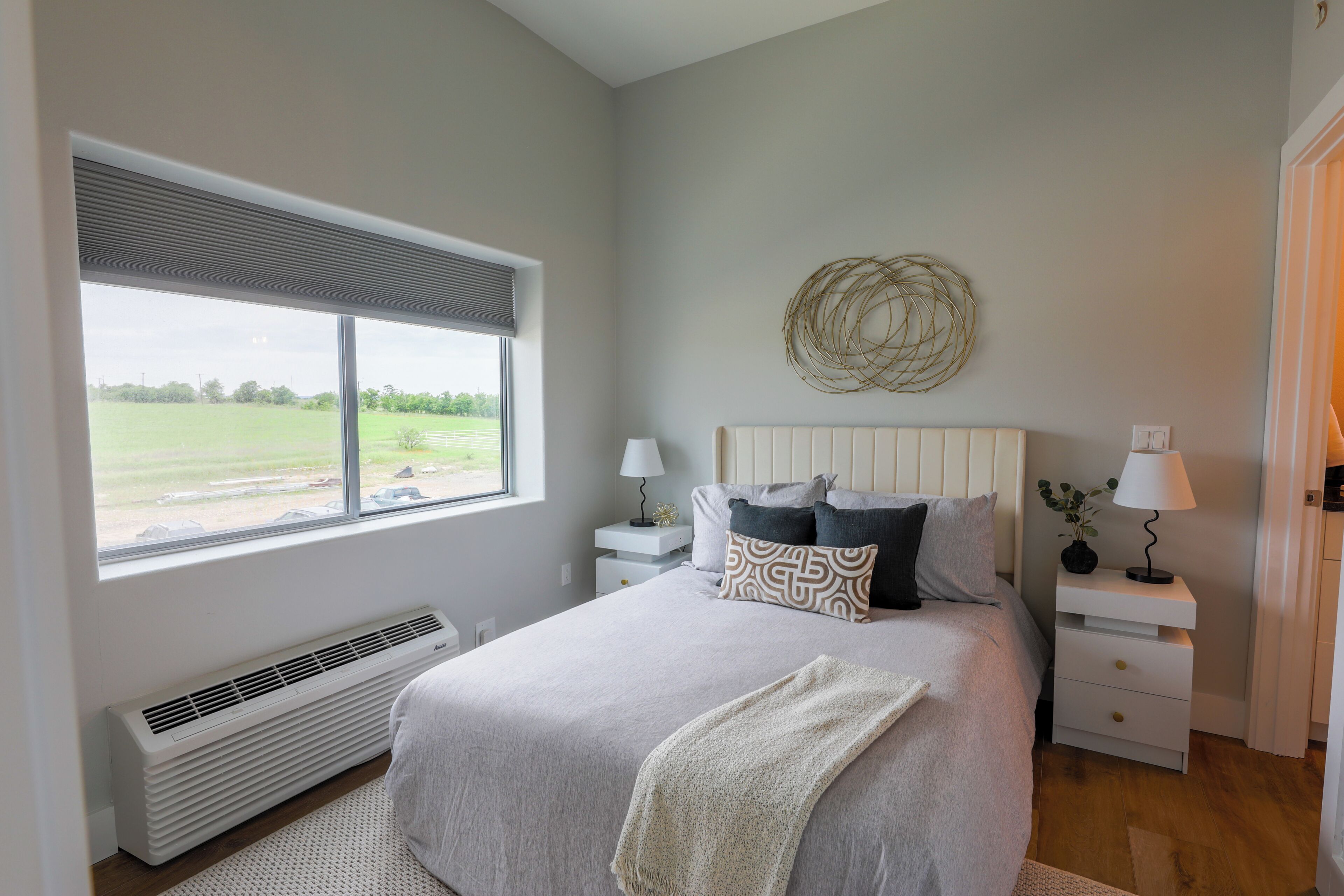 A bedroom with a large window, a bed with pillows, a bedside table with a lamp, and a wall-mounted decorative piece.