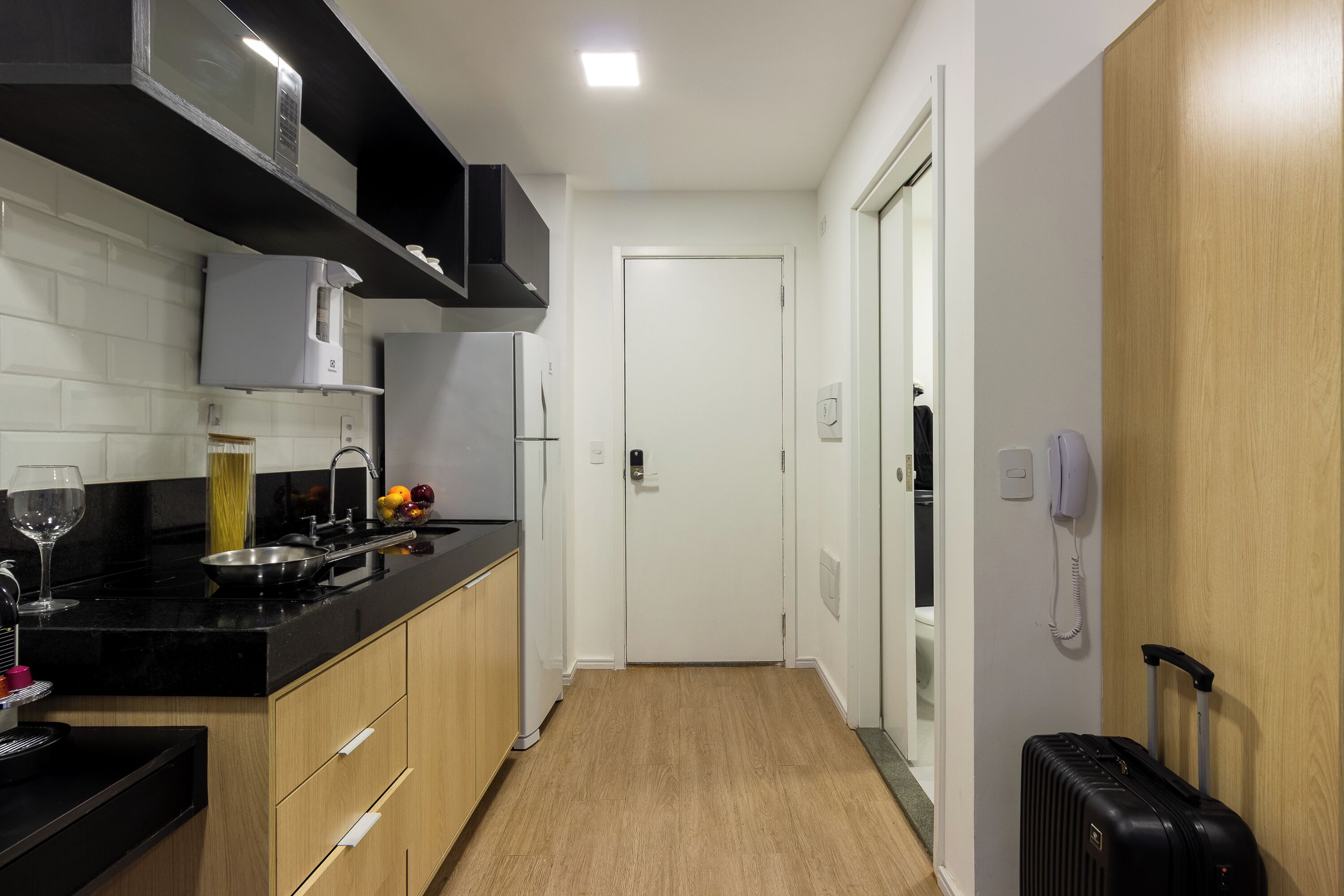 Een moderne keuken met zwarte aanrechtbladen, houten kasten en een zwarte koffer in de hal.