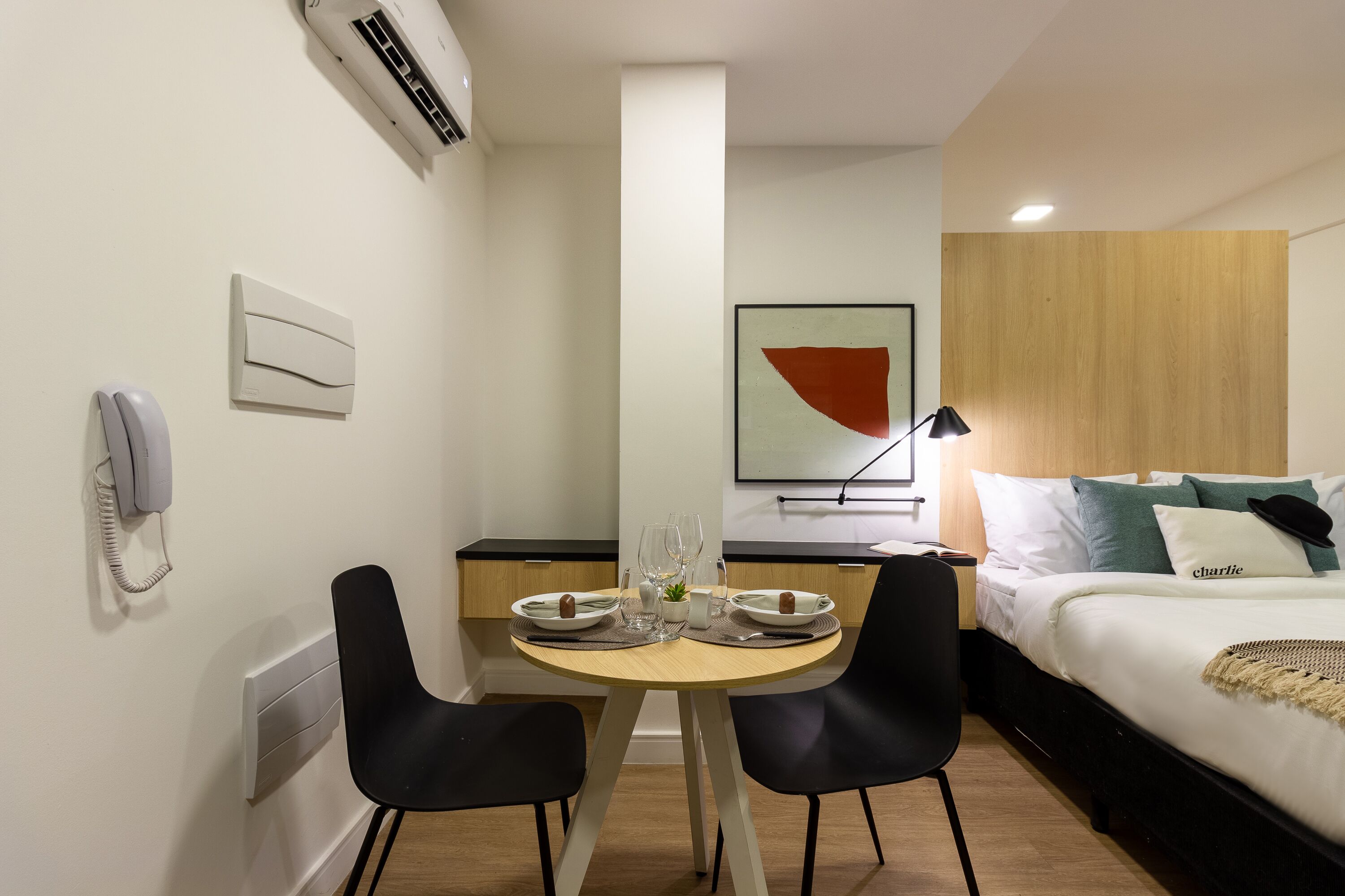 Ein Hotelzimmer mit einem Bett, einem Nachttisch mit Lampe, einem kleinen Tisch mit Stühlen, einem Telefon und einer Wandklimaanlage.