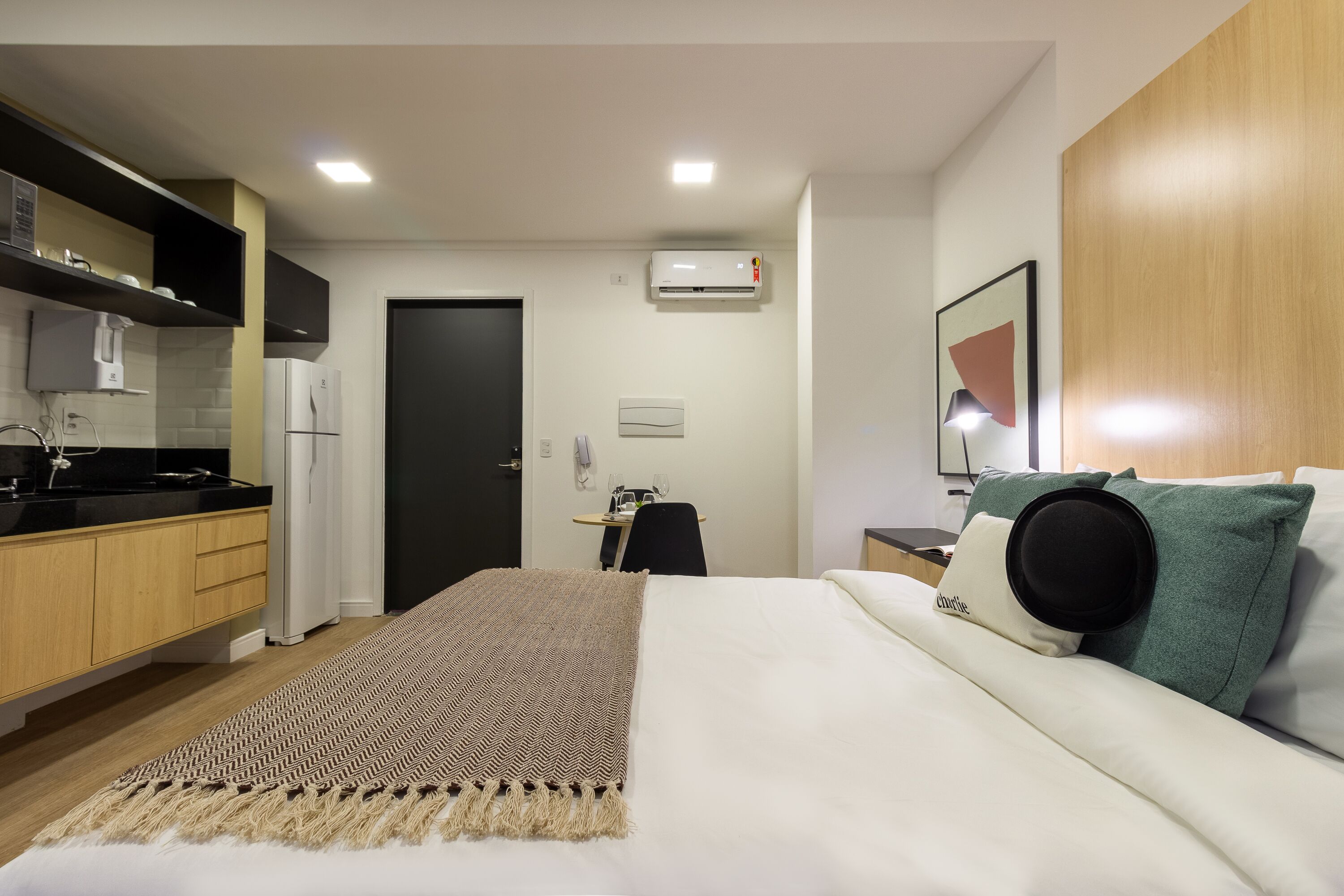 Ein modernes Hotelzimmer mit einer Küchenzeile, einem Bett mit Tagesdecke, einem Schreibtisch mit Stuhl und einer Wand-Klimaanlage.