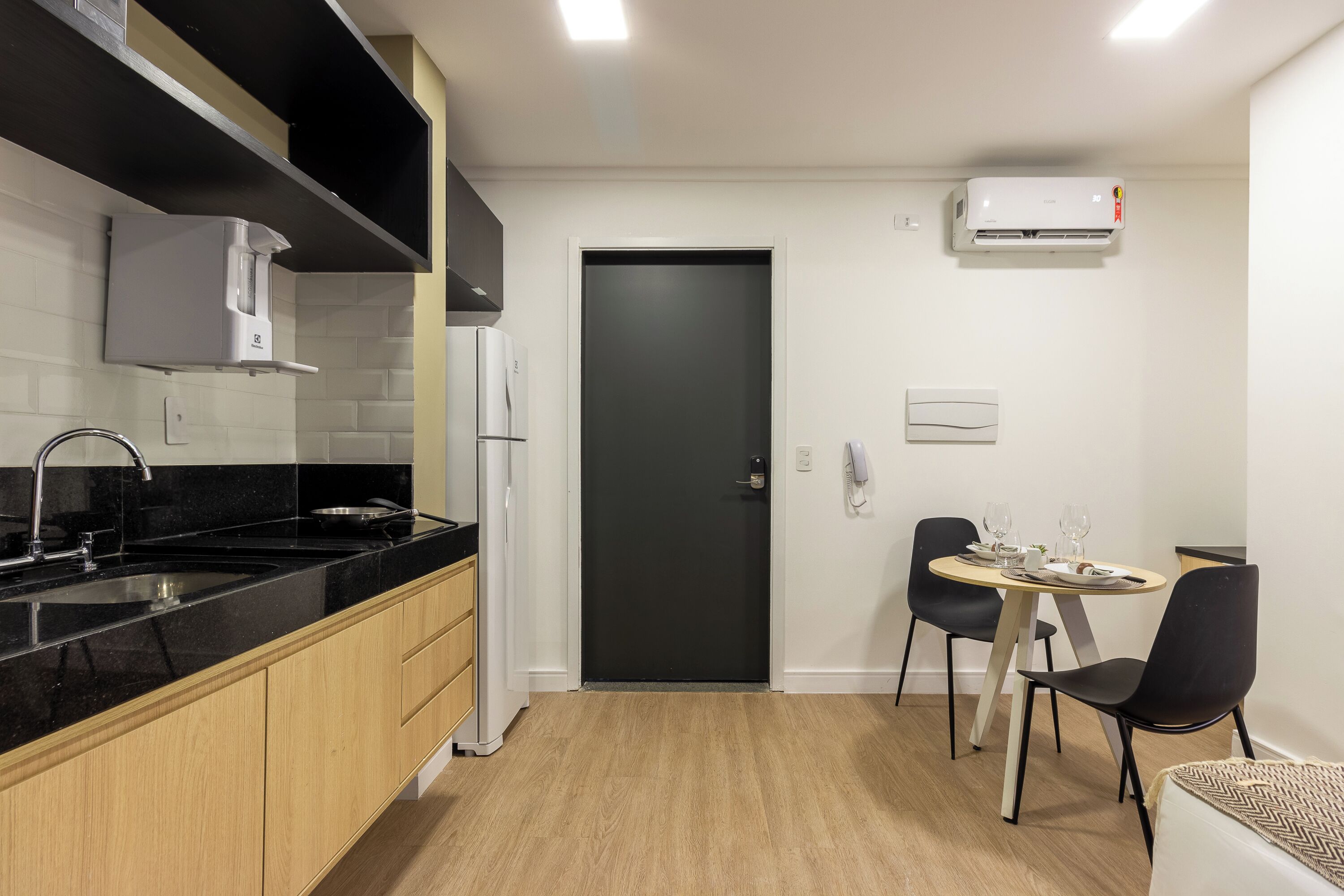 Una cocina moderna con encimera negra, armarios de madera, una puerta negra y una zona de comedor con mesa y sillas.