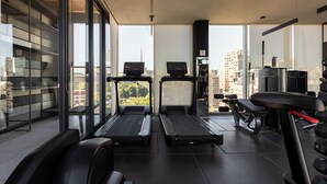 Ein modernes Fitnessstudio mit Laufbändern, Kraftgeräten und einer Bank. Große Fenster bieten Blick auf die Stadt.