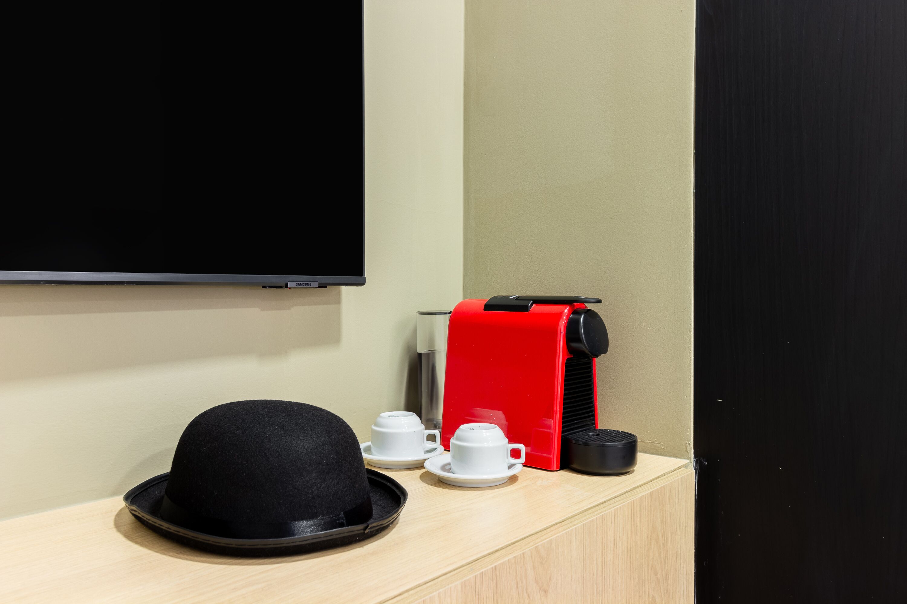 A hotel room with a television, a red coffee machine, a black hat, and two white cups on a wooden surface.