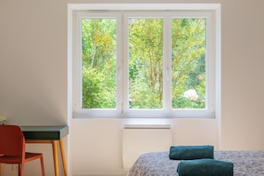 A room with a bed, a desk, and a window with a view of trees and flowers.