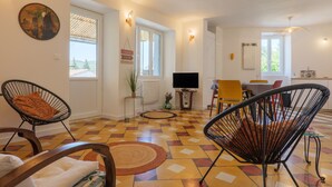 A living room with a TV, two chairs, a dining table, and a patterned floor. - Charming house in Vallon Pont d'Arc with private terrace, garden, and Wi-Fi (Vallon-Pont-d'Arc)