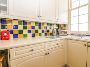 A kitchen with white cabinets, a colorful tile backsplash, a red first aid kit, a kettle, a toaster, and a window with a metal rack. - Lightkeepers Retreat (Eyemouth)