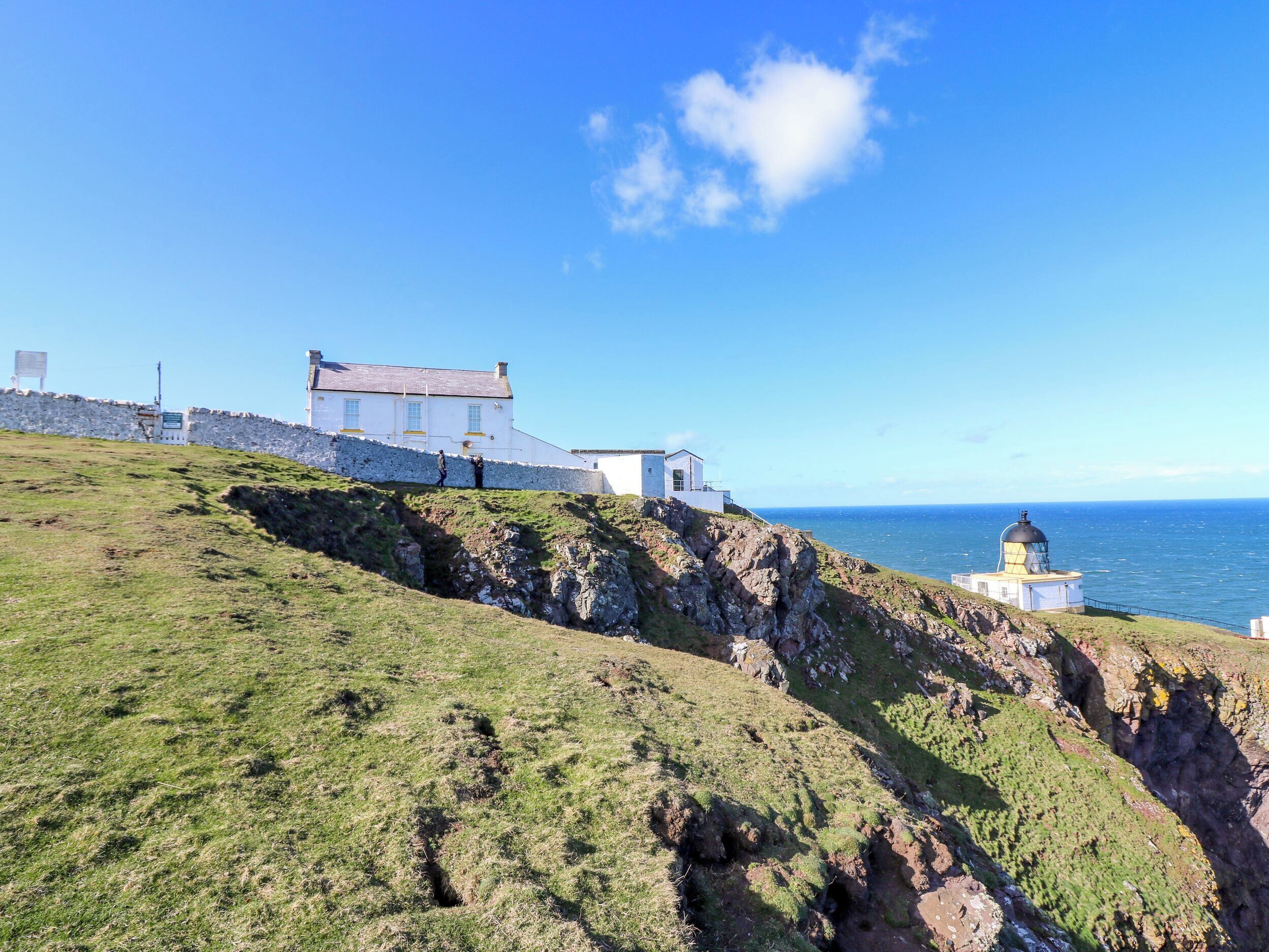A coastal building with a lighthouse, grassy terrain, and a stone wall.