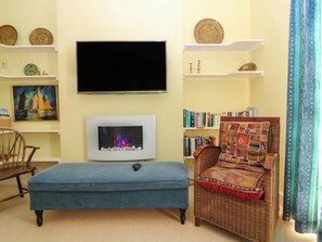 A living room with a flat-screen TV, a fireplace, a bookshelf, a wicker chair, and a blue ottoman. - Lightkeepers Retreat (Eyemouth)