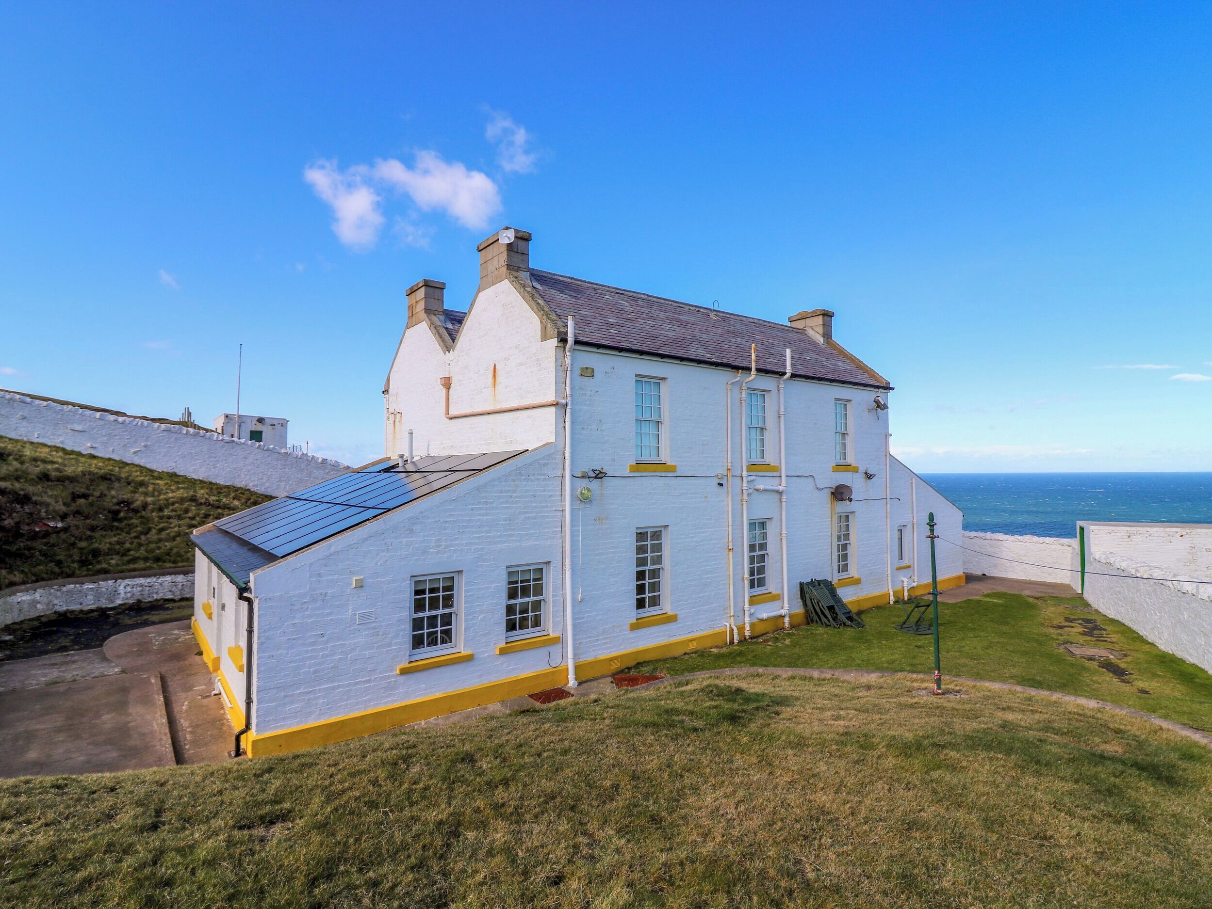 Un bâtiment blanc avec un toit en pente, une petite structure attenante dotée de panneaux solaires et une vue sur la mer.