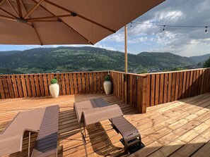Eine Holzterrasse mit zwei Liegestühlen und einem großen Sonnenschirm, mit Blick auf eine malerische Landschaft mit Hügeln und einem Dorf.