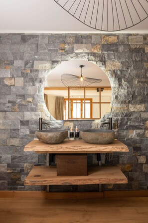 Un baño con pared de piedra, tocador de madera, espejo redondo y ventilador de techo.