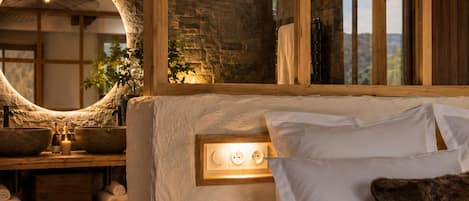 Un dormitorio con cama, cabecero de madera, espejo grande, pared de piedra y tragaluz circular.