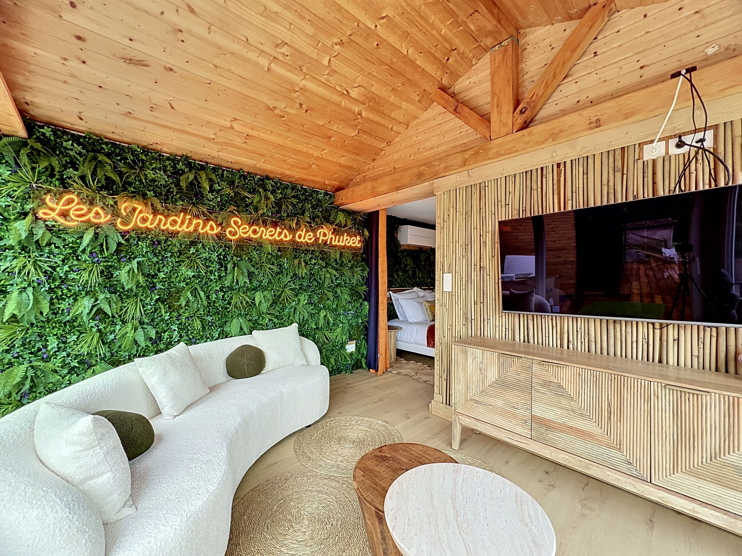 Une pièce avec un canapé blanc aux lignes courbes, un meuble TV en bois et un panneau néon sur lequel on peut lire : « Les Jardins Secrets de Phuket ».
