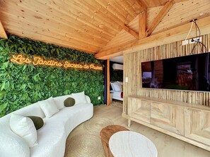 Une pièce avec un canapé blanc aux lignes courbes, un meuble TV en bois et un panneau néon sur lequel on peut lire : « Les Jardins Secrets de Phuket ».