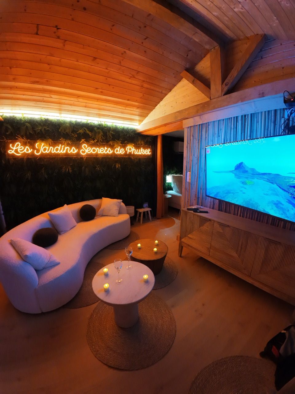 Un rincón lounge acogedor con un sofá blanco curvo, un televisor de pantalla plana y un letrero de neón que dice 'Les Jardins Secrets de Phuket'.