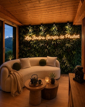 Une pièce chaleureuse avec un plafond en bois, un canapé avec des coussins, une table ronde en bois sur laquelle sont posés une théière et des tasses, et un panneau néon sur lequel on peut lire : « Les Jardins Secrets de Phuket ».