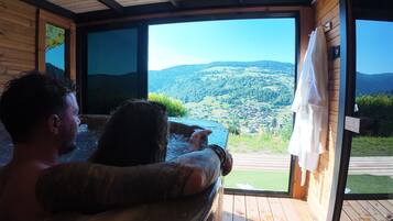 A person relaxing in a hot tub with a view of a mountainous landscape through a large window.
