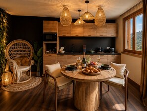 Un coin repas comprenant une table ronde en bois, deux chaises, un fauteuil en osier et une grande fenêtre donnant sur un paysage agréable.