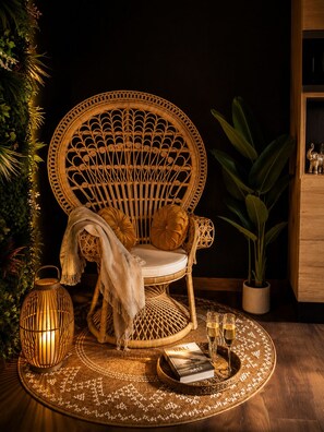 Une chaise en osier avec des coussins, une petite table avec un livre et des lunettes, une lanterne et une plante en pot.