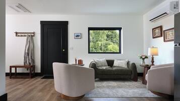 Una sala de estar con un sofá, dos sillones, una mesita de centro, una puerta, una ventana con vista a árboles y una lámpara.