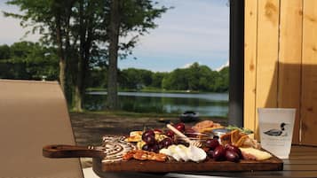 Una mesa con una tabla de quesos, frutas, frutos secos y una bebida, frente a un lago y árboles.