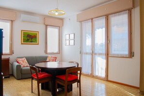 A dining area with a round table, chairs, a sofa, and a painting on the wall. - A cozy and comfortable 65 square meters apartment, perfect for five people, located on the first floor of a building without an elevator.The property enjoys an excellent location in Alghero, located on the street parallel to the seafront: you can reach A (Alghero)