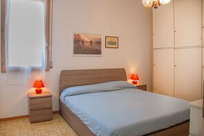 A bedroom with a bed, two bedside lamps, a window, a framed picture, and a wardrobe. - A cozy and comfortable 65 square meters apartment, perfect for five people, located on the first floor of a building without an elevator.The property enjoys an excellent location in Alghero, located on the street parallel to the seafront: you can reach A (Alghero)